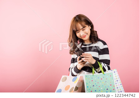 Portrait Asian happy beautiful young woman shopper smiling standing excited holding online shopping bags online multicolor and smartphone on hand, isolated on pink background, glad with mobile phone Portrait Asian happy beautiful young woman shopper smiling standing excited holding online shopping bags online multicolor and smartphone on hand, isolated on pink background, glad with mobile phone 122344742