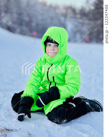 litte boy in neon green overall sitting on snow outside 122347488