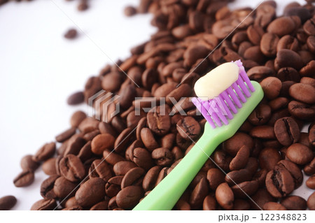A green toothbrush with purple bristles and a dollop of white toothpaste rests diagonally on a pile of roasted coffee beans, emphasizing the contrast between dental care and coffee culture. A green toothbrush with purple bristles and a dollop of white toothpaste rests diagonally on a pile of roasted coffee beans, emphasizing the contrast between dental care and coffee culture. 122348823