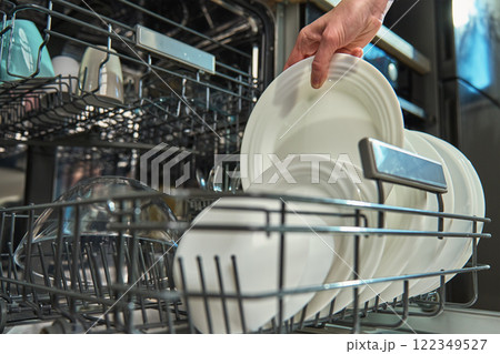 Woman unloading clean white plates from the bottom rack of dishwasher in kitchen. Person performing everyday household chores, using modern home appliance Woman unloading clean white plates from the bottom rack of dishwasher in kitchen. Person performing everyday household chores, using modern home appliance 122349527