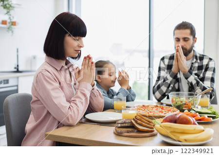 Caucasian family including mother, father, and daughter praying before breakfast. Seen in modern kitchen, focused on faith and gratitude. Represents harmony and family bonding. Caucasian family including mother, father, and daughter praying before breakfast. Seen in modern kitchen, focused on faith and gratitude. Represents harmony and family bonding. 122349921