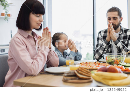 Caucasian family with parents and daughter praying together before breakfast. Meal on table includes fruit, pizza, and salad. Moment of gratitude and connection. Caucasian family with parents and daughter praying together before breakfast. Meal on table includes fruit, pizza, and salad. Moment of gratitude and connection. 122349922