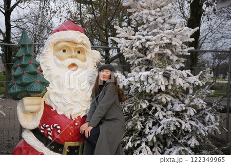 Woman posing with giant Santa Claus statue and snowy Christmas tree outdoors. Woman posing with giant Santa Claus statue and snowy Christmas tree outdoors. 122349965