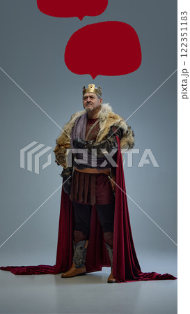Art collage. Medieval king in golden crown, fur-lined cloak, and armor standing on grey background surrounded by red speech bubbles for text placement. 122351183