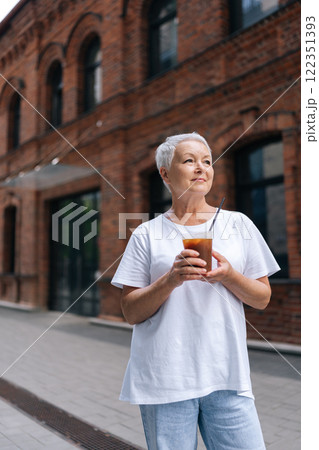 Vertical portrait of relaxed senior woman with stylish short haircut, dressed in casual attire, savoring an iced coffee while standing on city sidewalk, enjoying free time in retirement outdoors. Vertical portrait of relaxed senior woman with stylish short haircut, dressed in casual attire, savoring an iced coffee while standing on city sidewalk, enjoying free time in retirement outdoors. 122351393