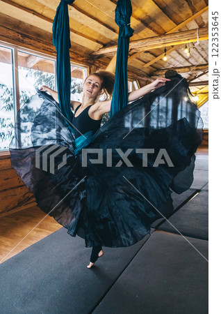 Girl gymnast in black dress doing aerial yoga. Fly yoga in hammock in spacious hall of wooden house Girl gymnast in black dress doing aerial yoga. Fly yoga in hammock in spacious hall of wooden house 122353645