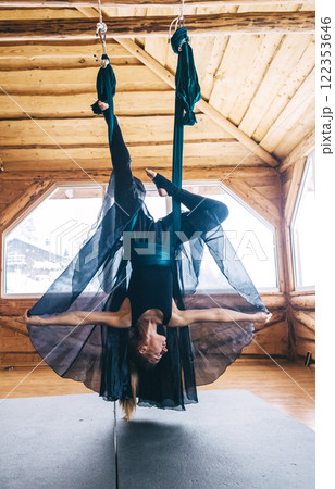 Girl gymnast in black dress doing aerial yoga. Fly yoga in hammock in spacious hall of wooden house Girl gymnast in black dress doing aerial yoga. Fly yoga in hammock in spacious hall of wooden house 122353646