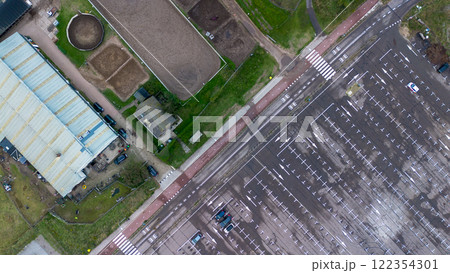 Aerial view of an empty parking lot, a large building, and equestrian facilities with paddocks, surrounded by green grass and pathways, captured from above. Aerial view of an empty parking lot, a large building, and equestrian facilities with paddocks, surrounded by green grass and pathways, captured from above. 122354301
