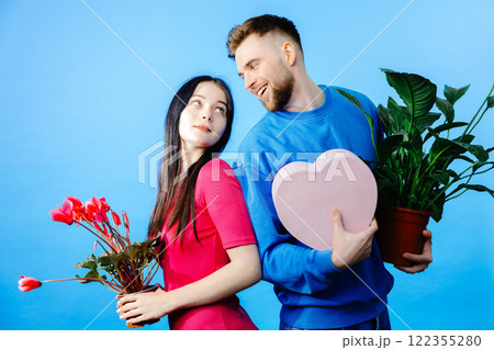 Loving boyfriend holding flowers pot and box and looking to girlfriend with present making romantic surprise Loving boyfriend holding flowers pot and box and looking to girlfriend with present making romantic surprise 122355280
