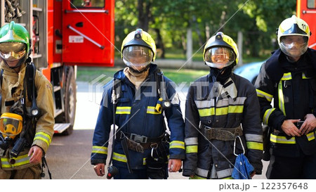 Portrait of male and female firefighters in helmets and protective uniforms standing near big red car. Young confident fireman and firewoman in full equipment posing against background of fire engine 122357648