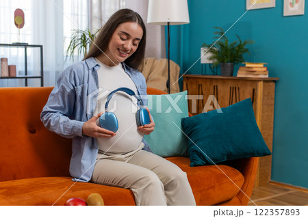 Pregnant woman sitting on home sofa listening music putting headphones on her tummy with child baby 122357893