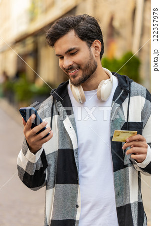 Smiling young man using credit card smartphone while transferring money purchases online outdoors Smiling young man using credit card smartphone while transferring money purchases online outdoors 122357978