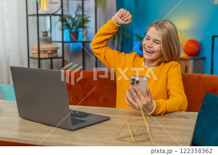 Teenager boy winning virtual school competition on laptop, celebrating exam success with smartphone 122358362