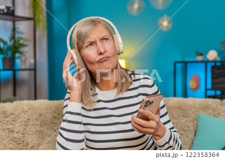 Mature woman listening to music on smartphone with wireless headphones dancing resting on home sofa Mature woman listening to music on smartphone with wireless headphones dancing resting on home sofa 122358364