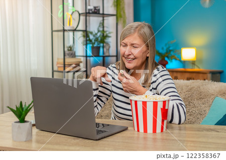 Mature woman watching movie on laptop with popcorn, laughing and enjoying content on sofa at home Mature woman watching movie on laptop with popcorn, laughing and enjoying content on sofa at home 122358367
