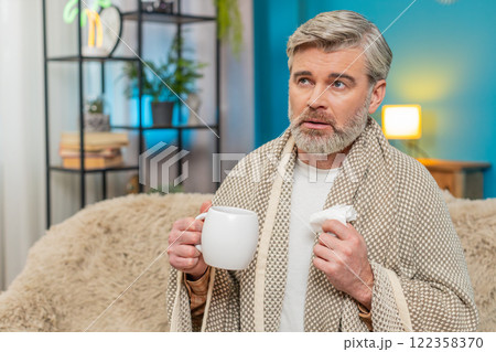 Mature man sitting at home holding napkin wipes snot drinking hot beverage or cold medicine at home 122358370