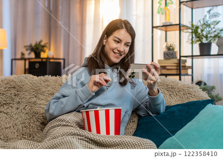 Happy Caucasian relaxed woman girl sitting at home sofa watching series on smartphone eating popcorn Happy Caucasian relaxed woman girl sitting at home sofa watching series on smartphone eating popcorn 122358410