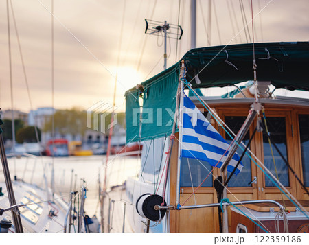 View of Greek flag waving in air serene Mediterranean vibe. beautiful Greek islands series, sailing yacht in harbour offering tourists daily cruises along side Greek flag. 122359186