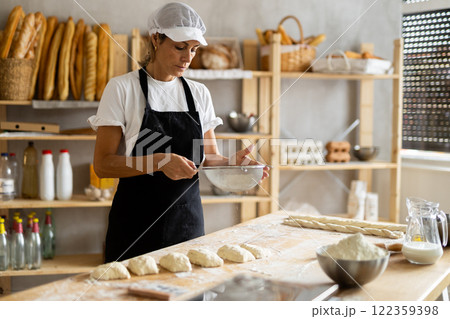 Working in bakery - female baker sprinkle flour onto dough through sieve 122359398