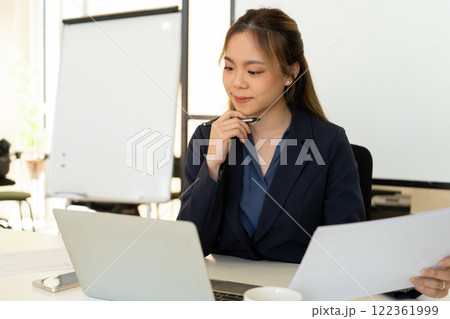 Businesswoman sitting at desk on couch in workplace or at home working on laptop and analyzing data on charts and graphs and writing on papers to make business plan and strategies for company. Businesswoman sitting at desk on couch in workplace or at home working on laptop and analyzing data on charts and graphs and writing on papers to make business plan and strategies for company. 122361999