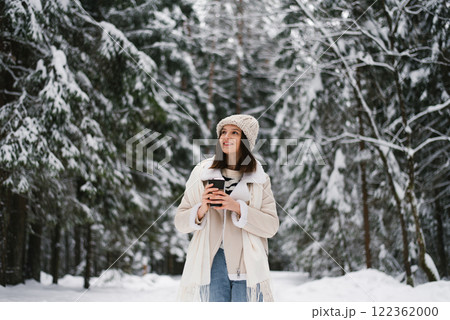 Smiling woman wearing white coat, hat is standing in snowy forest and holding cup of coffee Smiling woman wearing white coat, hat is standing in snowy forest and holding cup of coffee 122362000