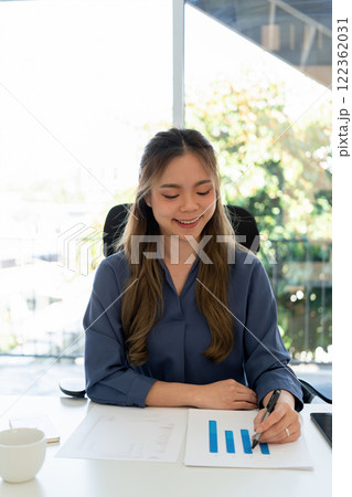 Businesswoman sitting at desk on couch in workplace or at home working on laptop and analyzing data on charts and graphs and writing on papers to make business plan and strategies for company. Businesswoman sitting at desk on couch in workplace or at home working on laptop and analyzing data on charts and graphs and writing on papers to make business plan and strategies for company. 122362031