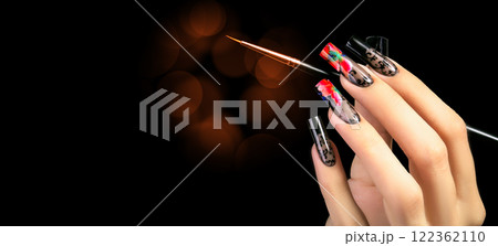 A hand with a beautiful manicure and a brush on a black background with burgundy bokeh spots. Nail design. Extended nails. 122362110