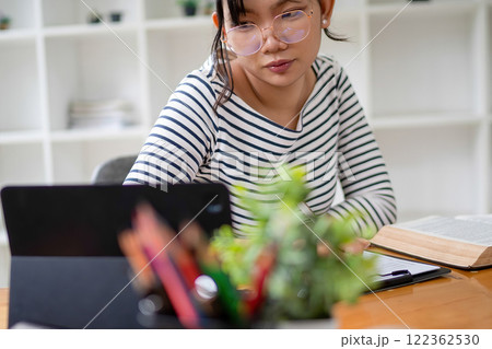 Intelligent young woman having online lessons on laptop, drinking coffee and taking notes, 122362530