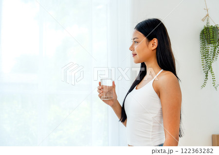 Close up and side view of beautiful Asian woman hold glass of water and stand in front of glass window with day light and she look happy with look at the glass. Close up and side view of beautiful Asian woman hold glass of water and stand in front of glass window with day light and she look happy with look at the glass. 122363282