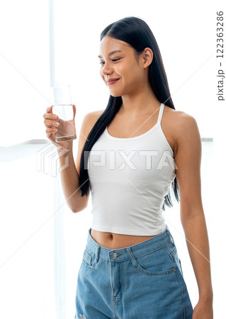 Vertical image of beautiful Asian woman hold glass of water and stand in front of glass window with day light and she look happy with look at the glass. Vertical image of beautiful Asian woman hold glass of water and stand in front of glass window with day light and she look happy with look at the glass. 122363286