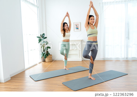 Side view couple Asian women exercise together with yoga posture by stand and hands together over head also stay on mat in room with day light. Side view couple Asian women exercise together with yoga posture by stand and hands together over head also stay on mat in room with day light. 122363296