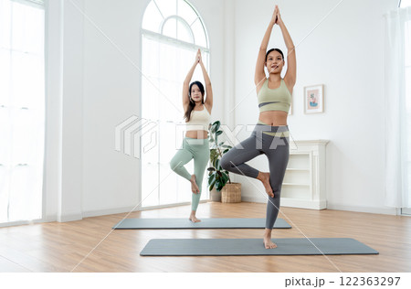 Couple Asian women exercise together with yoga posture by stand and hands together over head also stay on mat in room with day light. Couple Asian women exercise together with yoga posture by stand and hands together over head also stay on mat in room with day light. 122363297