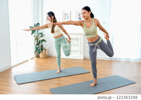 Wide shot Asian women exercise together with yoga posture by stand with raise right arm forward hold left leg to back on mat in room with day light. Wide shot Asian women exercise together with yoga posture by stand with raise right arm forward hold left leg to back on mat in room with day light. 122363298