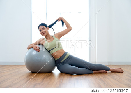 Beautiful Asian woman with exercise clothes lie on exercise grey ball with use right hand lean to ball and use left hand hold her hair and look at camera with day light in front of glass window. Beautiful Asian woman with exercise clothes lie on exercise grey ball with use right hand lean to ball and use left hand hold her hair and look at camera with day light in front of glass window. 122363307