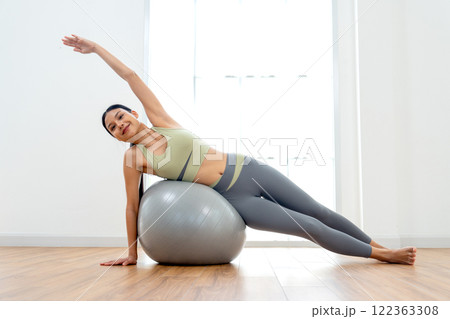 Beautiful Asian woman with exercise clothes lie on exercise grey ball with use right hand push up and raise left hand up over her head and look at camera with day light in front of glass window. Beautiful Asian woman with exercise clothes lie on exercise grey ball with use right hand push up and raise left hand up over her head and look at camera with day light in front of glass window. 122363308
