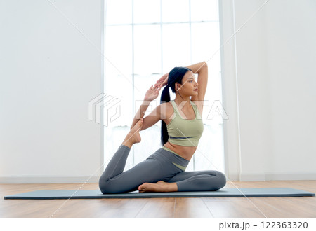 Pretty Asian woman with exercise clothes sit and do yoga exercise posture by hold both hand and rest foot up to touch elbow with turn face to her left side and stay in front of glass window. Pretty Asian woman with exercise clothes sit and do yoga exercise posture by hold both hand and rest foot up to touch elbow with turn face to her left side and stay in front of glass window. 122363320