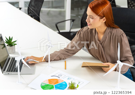 Young female engineer sitting at the table and looking at the wind turbine model Young female engineer sitting at the table and looking at the wind turbine model 122365394