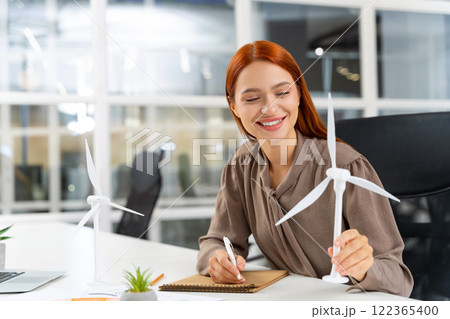 Young female engineer sitting at the table and looking at the wind turbine model 122365400