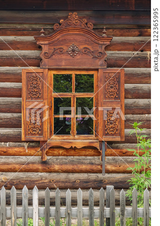 Ornate wooden window frame adorning traditional russian log cabin, framing verdant landscape and highlighting intricate woodworking artistry 122365995