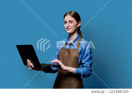 Portrait of young woman in apron holding laptop on blue background 122366103