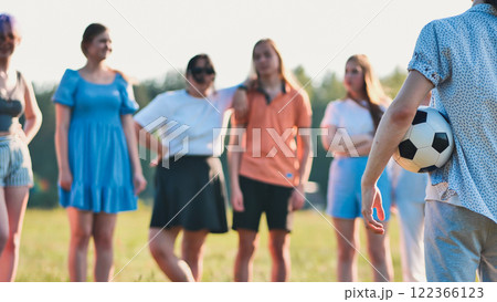 Group of friends in a field watches as a young man walks away with a soccer ball 122366123