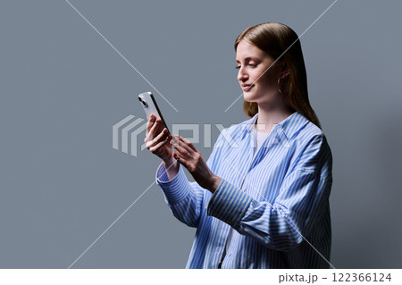 Teenage student girl in blue shirt holding smartphone in hands on grey studio background Teenage student girl in blue shirt holding smartphone in hands on grey studio background 122366124