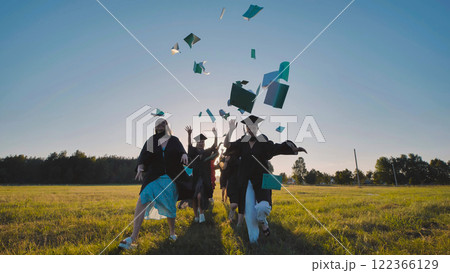 Happy graduates celebrating their academic success by tossing books into the air while running in a field wearing academic gowns and caps Happy graduates celebrating their academic success by tossing books into the air while running in a field wearing academic gowns and caps 122366129