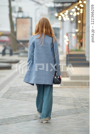 Back view of a young woman walking down a bustling city street in early autumn, dressed in a blue coat and oversized jeans 122366209