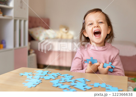smiling very excited little child girl connecting puzzle pieces on wooden desk in room smiling very excited little child girl connecting puzzle pieces on wooden desk in room 122366638