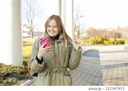 Smiling woman using mobile phone app, playing game, shopping online outdoors. Happy woman celebrating victory in lottery. Amazed hipster wondered with win in online auction betting on website Smiling woman using mobile phone app, playing game, shopping online outdoors. Happy woman celebrating victory in lottery. Amazed hipster wondered with win in online auction betting on website 122367451