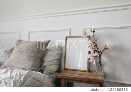 Blank wooden picture frame mockup on wooden side table with glass vase. Blooming magnolia tree branches. Scandinavian interior. Elegant bedroom, beige pillows. White wall background, stucco decor. Blank wooden picture frame mockup on wooden side table with glass vase. Blooming magnolia tree branches. Scandinavian interior. Elegant bedroom, beige pillows. White wall background, stucco decor. 122368558