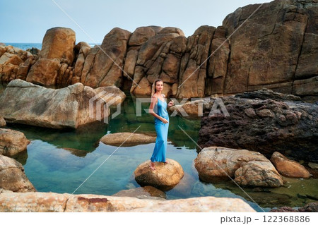 Stunning model in wet blue dress stands on rock at exotic ocean beach. Luxury fashion photoshoot. Natural water pool and rocky landscape. Perfect for high-end beauty, fragrance, travel ads. 122368886