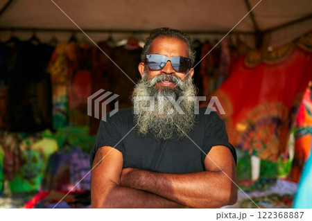 Senior artist with big beard, sunglasses stands in front of colorful tie-dye t-shirts at vibrant fair. Bearded man crosses arms, smiles confidently. Artisan spirit in open-air market. Senior artist with big beard, sunglasses stands in front of colorful tie-dye t-shirts at vibrant fair. Bearded man crosses arms, smiles confidently. Artisan spirit in open-air market. 122368887