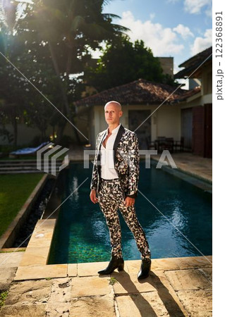 Bald man in bold floral suit by villa pool. Stylish gay groom ready for wedding, sunlit luxury villa setting. Resort fashion, chic outfit for special occasion. Bald man in bold floral suit by villa pool. Stylish gay groom ready for wedding, sunlit luxury villa setting. Resort fashion, chic outfit for special occasion. 122368891